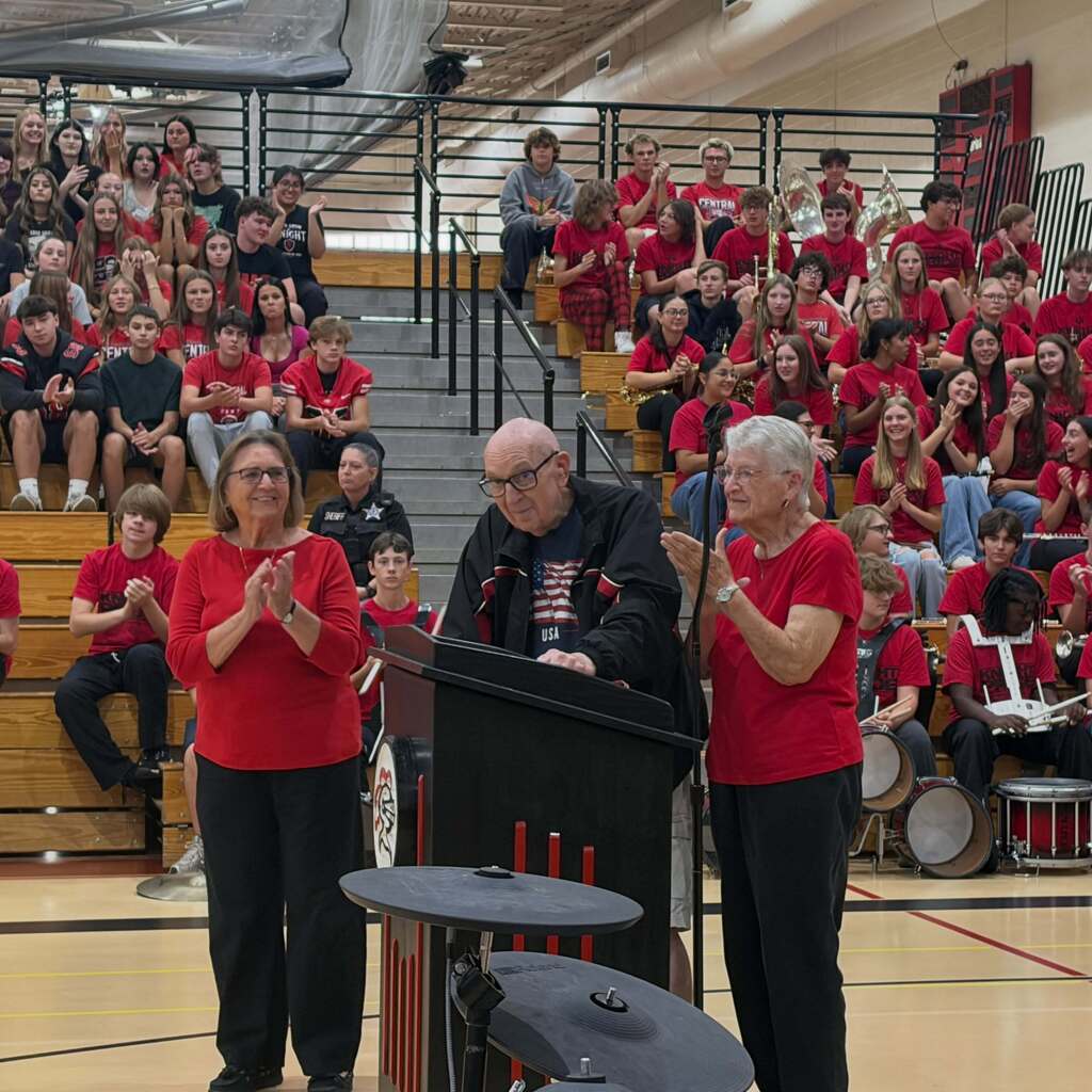 Three people in red shirts stand at a podium in a gymnasium. Behind them, a crowd sits on bleachers.