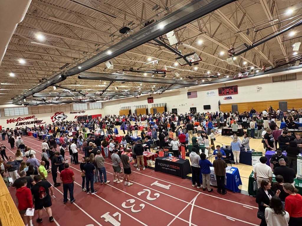 Large indoor area with people, tables, and a running track. Many people are gathered, some wearing sneakers.