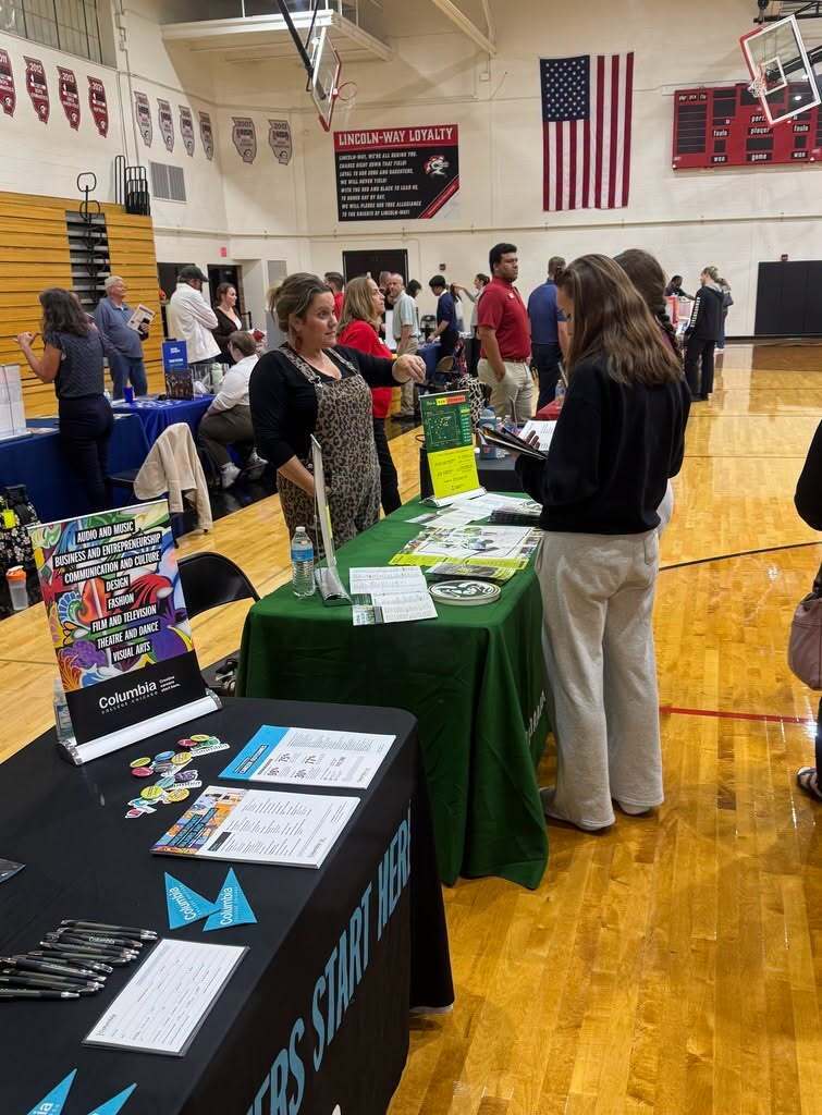 A group of people inside a gym with wooden floors. Two women stand behind a table with pamphlets and items.