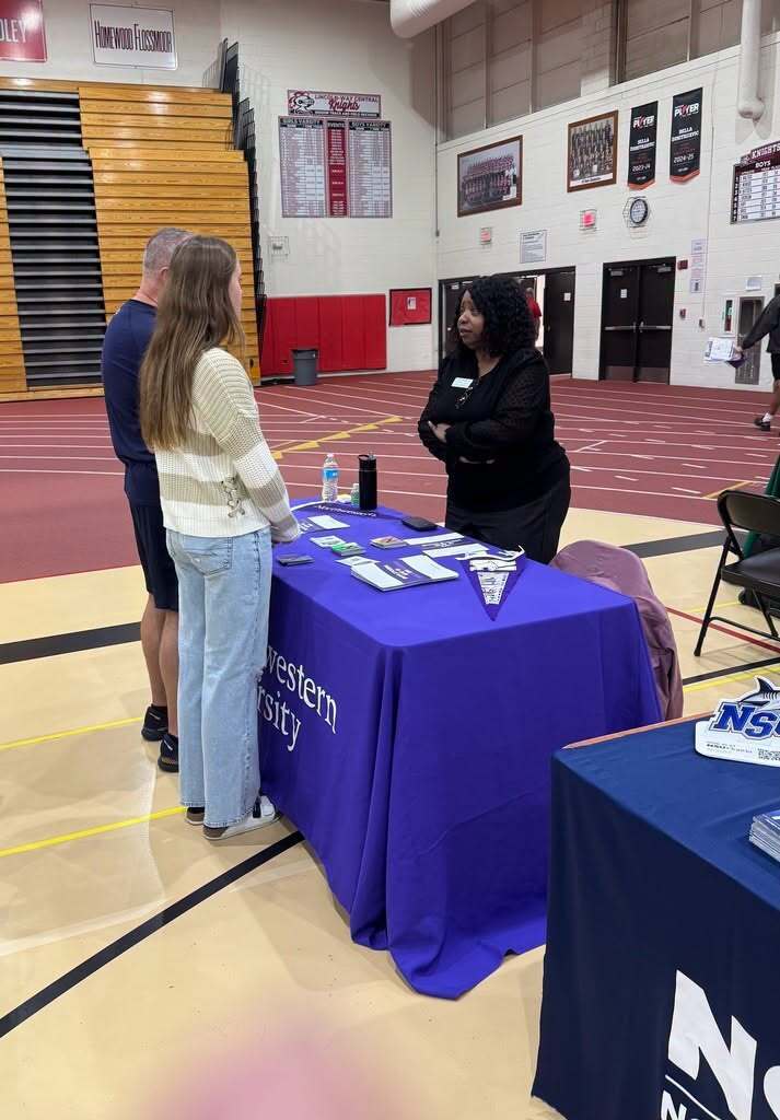 Two women standing at a table with a blue cover in a gymnasium with a track.