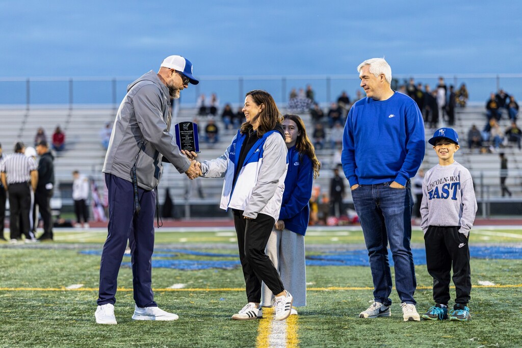 People stand on a field. A man presents an award to a woman. Spectators watch from bleachers.