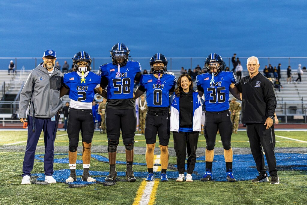 Five athletes and two coaches stand on a football field at dusk. The athletes wear blue uniforms with numbers 58, 0, and 36. Coaches wear black.