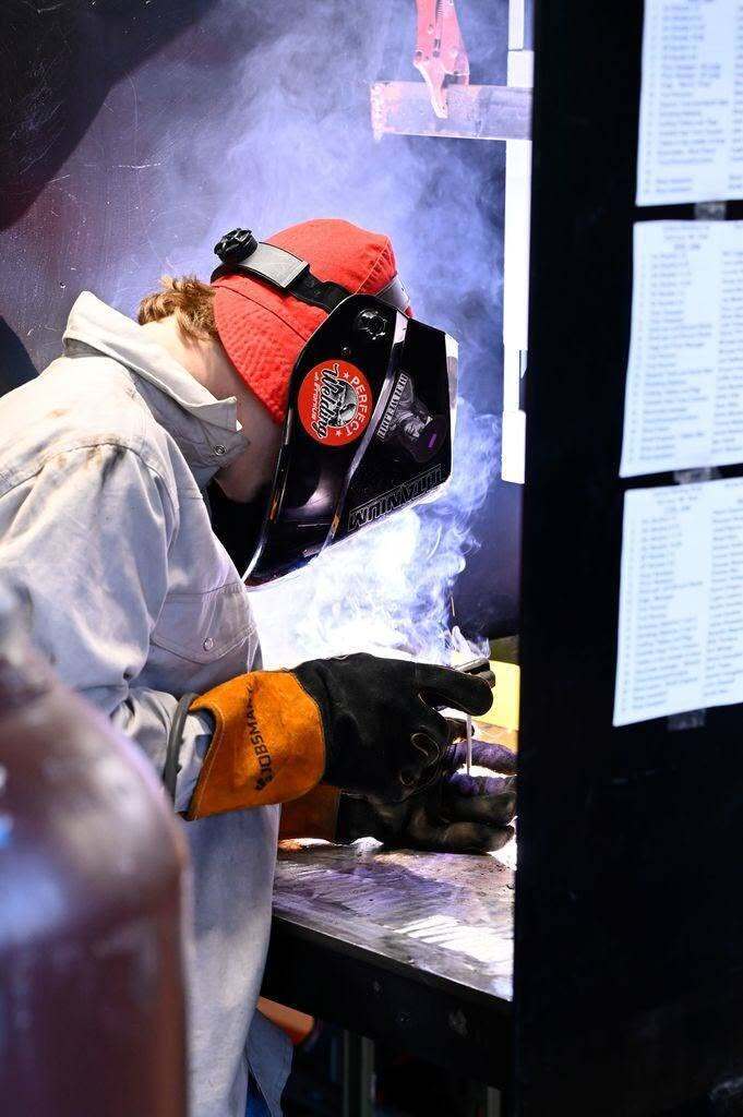 A person in a welding mask, red hat, and gloves works on a metal surface.