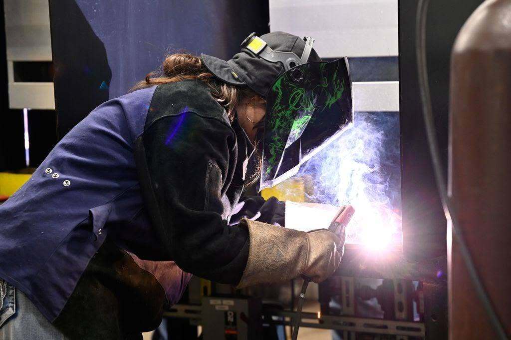 A person in a welding mask welds metal. The scene is well-lit, with shadows in the background.