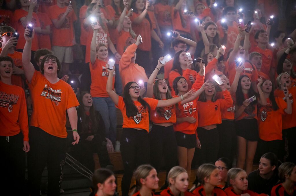 Group of people in orange shirts, holding up lit phones, cheering at a sports event.