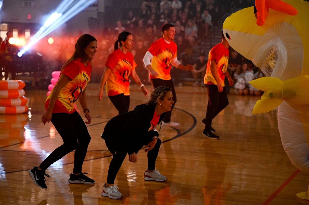 People in bright shirts dance on a court, led by a person with a microphone. A large, yellow mascot joins them.