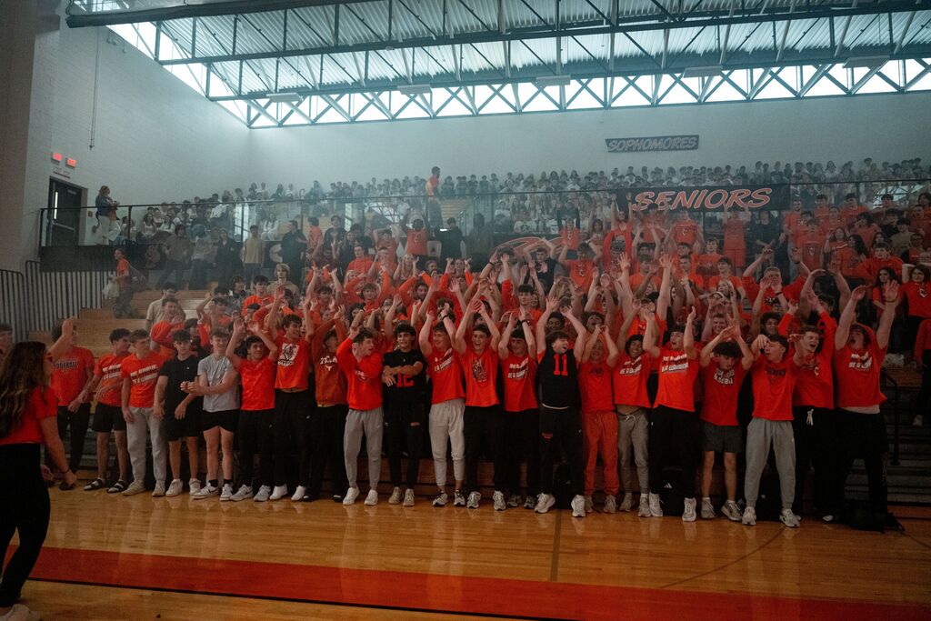 A group of people in orange shirts and sneakers stand on a court, raising hands in a gymnasium.