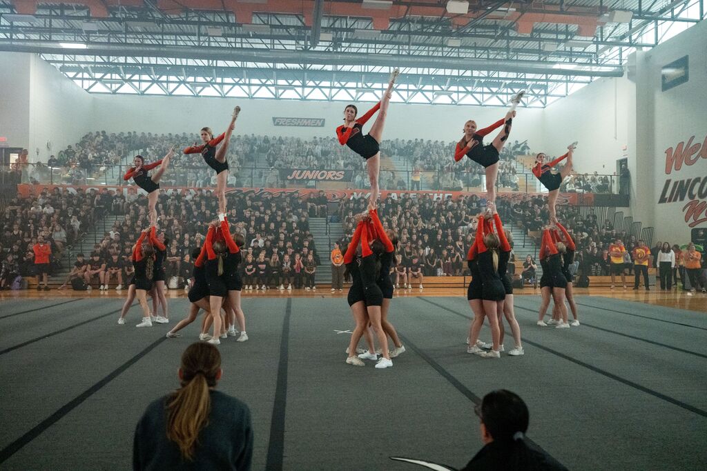 Group of cheerleaders performing stunts on a stage with a crowd watching from the stands.