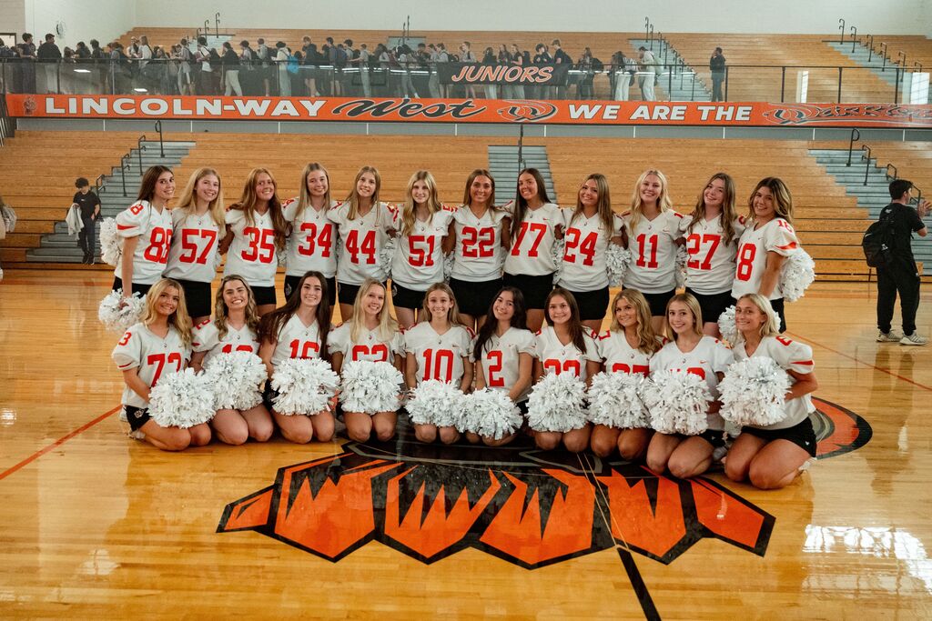 Team of cheerleaders in white tops and skirts pose on a basketball court in front of an orange lion logo.