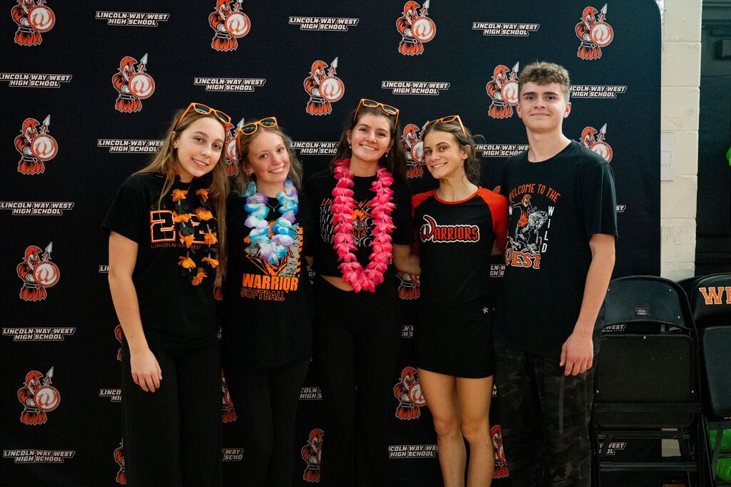 Four individuals stand together against a decorative wall, wearing matching black shirts with team-themed graphics. One woman wears a lei, while another wears sunglasses on her head.