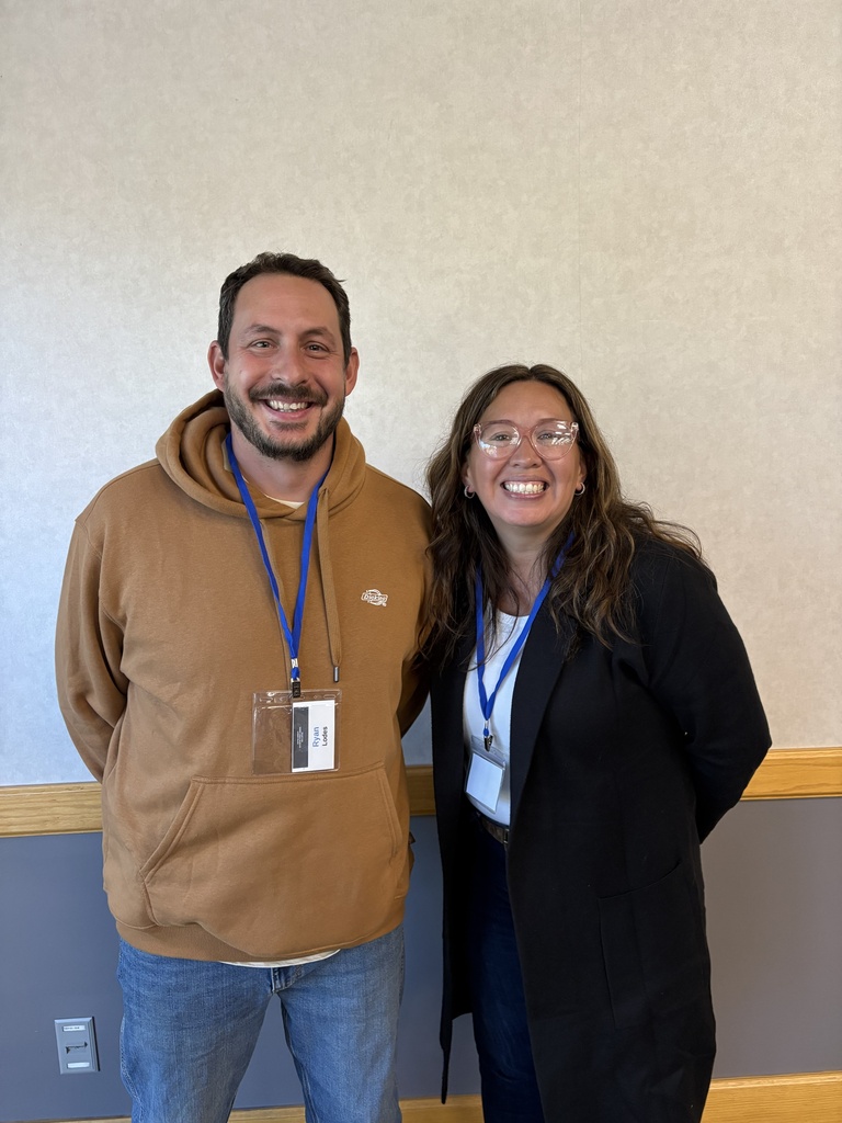 Two teachers smiling and posing for a photo against a gray wall