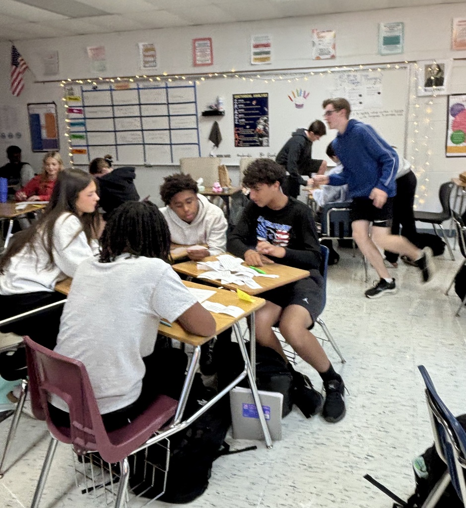 Students seated at desks in a classroom, engaged in activities. A person runs past them.