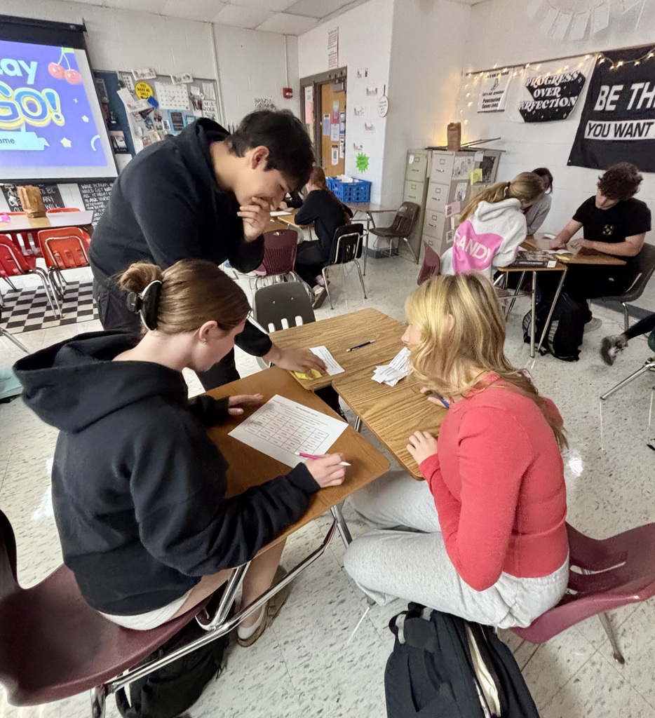 Students in a classroom, some seated at desks and others standing, engage in writing activities.