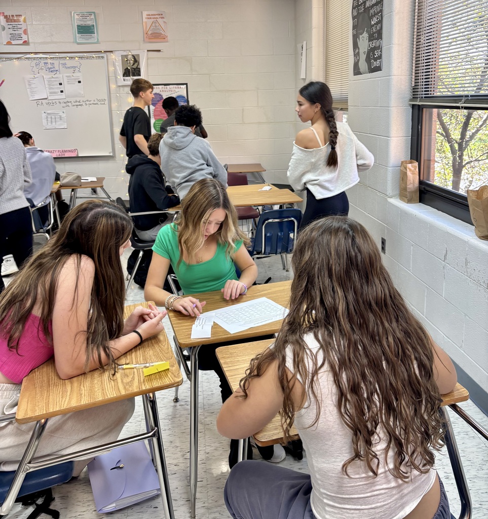 A classroom with students seated at desks, some engaged in work. A whiteboard with writing, a window with a view of trees, and a person standing.