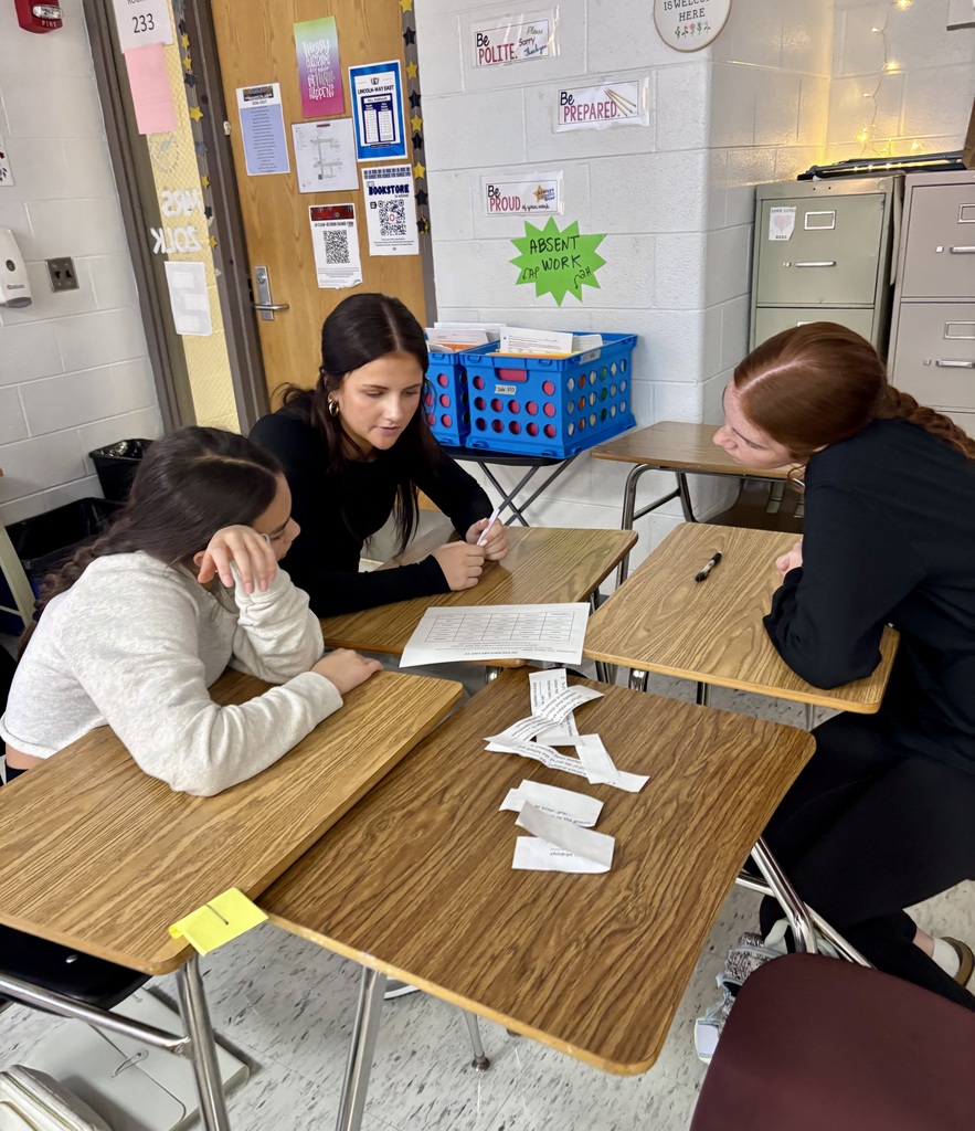 Three students seated at desks with papers and pens in a classroom setting.