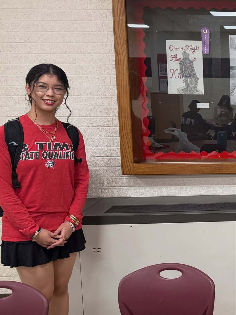 Student in a red top and skirt stands by chairs and a display case.