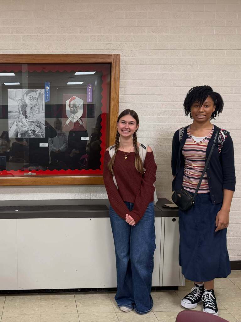 Two students stand near a wall with a framed art display behind them. One has braids, a backpack, and wears a maroon shirt. The other has a striped shirt, a bag, and blue skirt.
