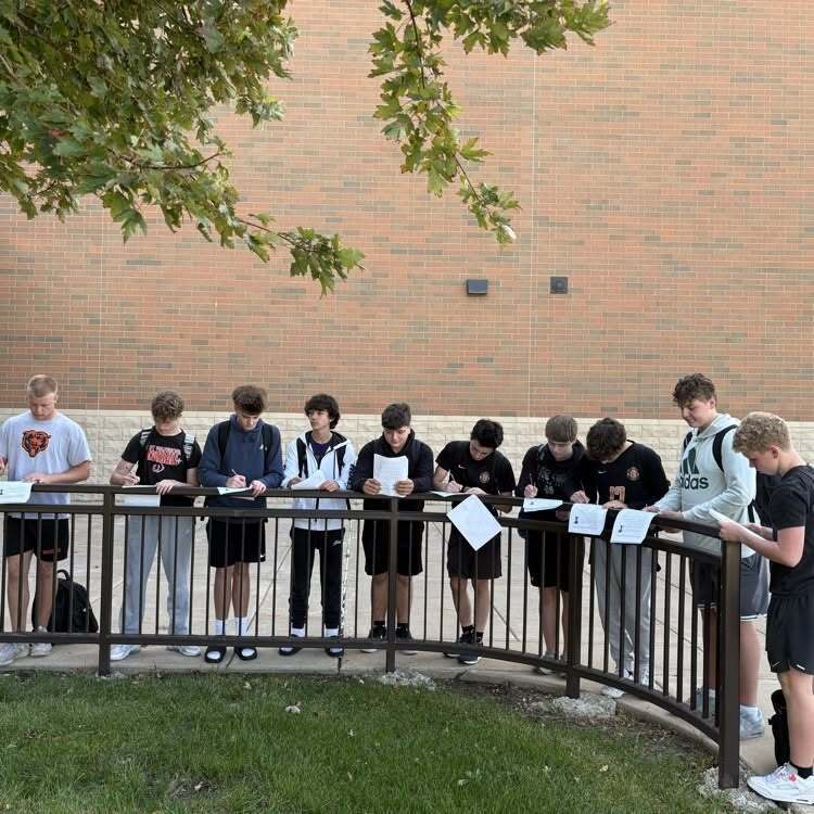 Students standing behind a fence, holding papers, in front of a brick building.