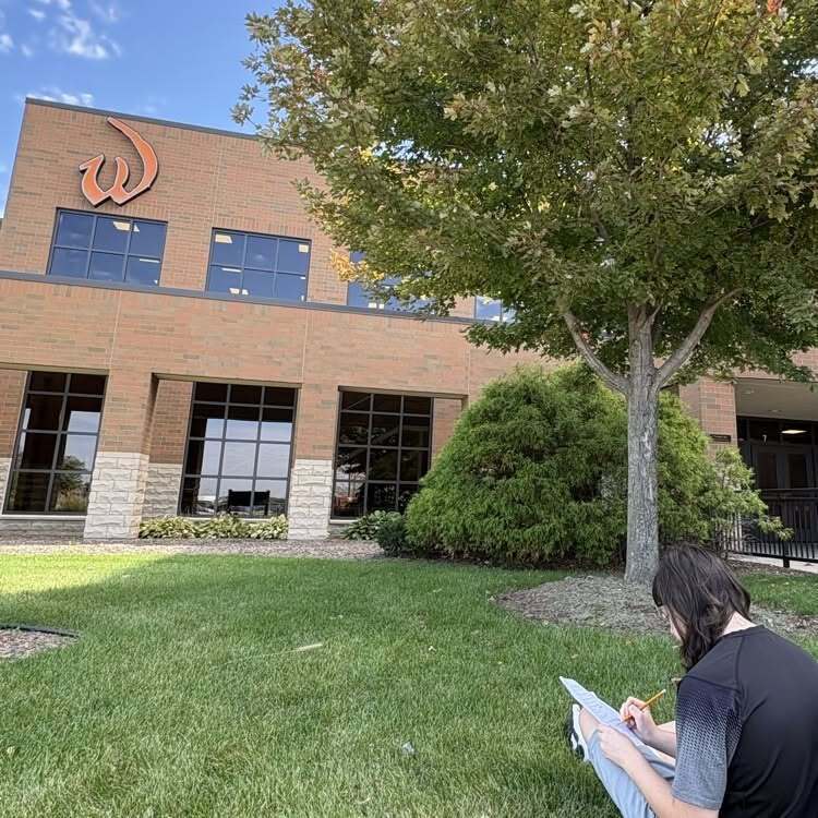 Person seated in front of a tree, writing. A large building with a logo on the side is behind.