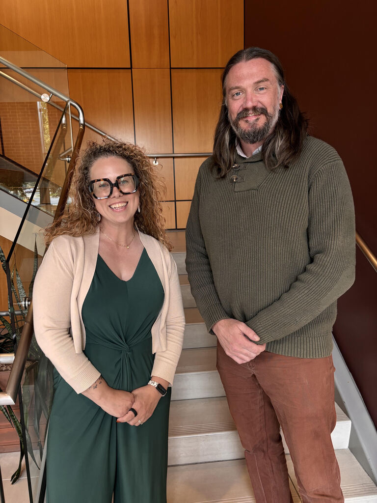 Two adults stand near a staircase. The woman wears a green dress and glasses. The man wears a green sweater and brown pants.