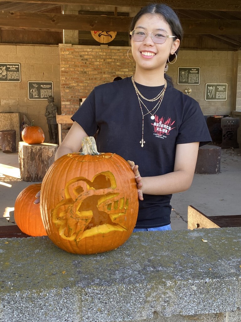 Girl smiling, holding pumpkin with a carved face in front of other pumpkins and stone barrier.