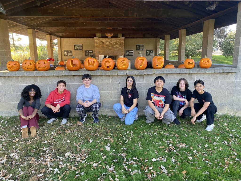 Eight students sit in front of a wall with carved pumpkins in numbers and letters.