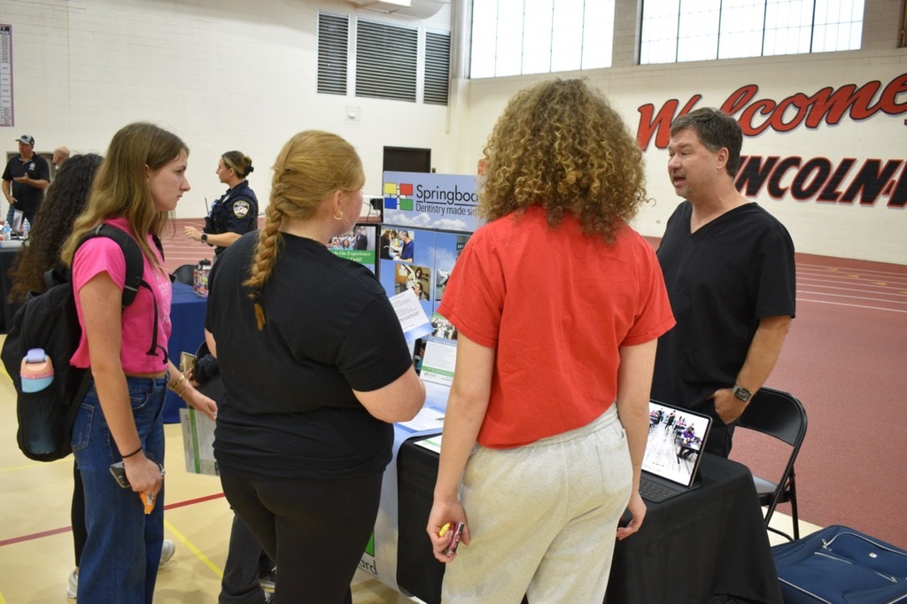 Five people in a gymnasium talk around a table with informational posters.