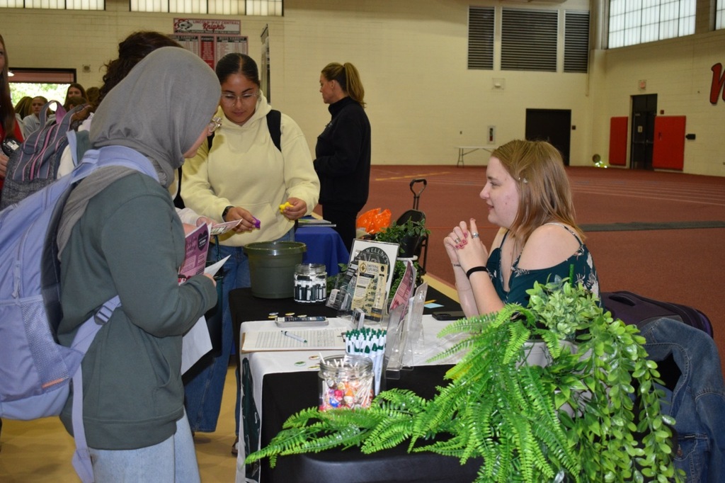 People at a table with a plant, jars, and other objects; two women look at each other.