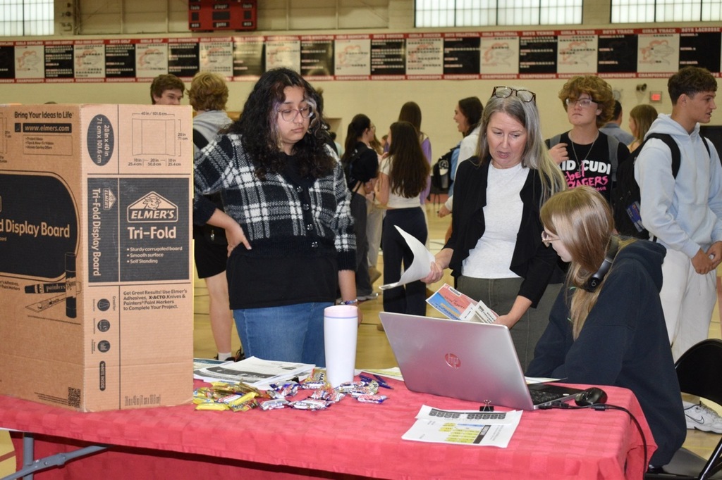 Indoors, a group of people stand around a table with a laptop, papers, and a box.