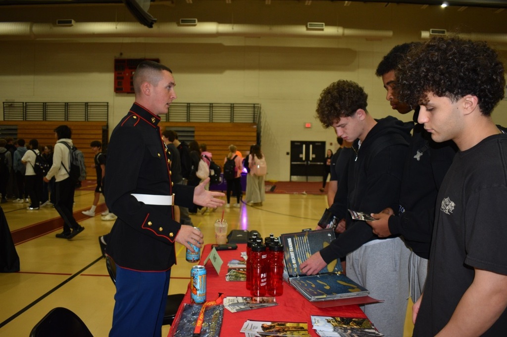 A uniformed military representative speaks with a group of students around a table displaying various pamphlets.