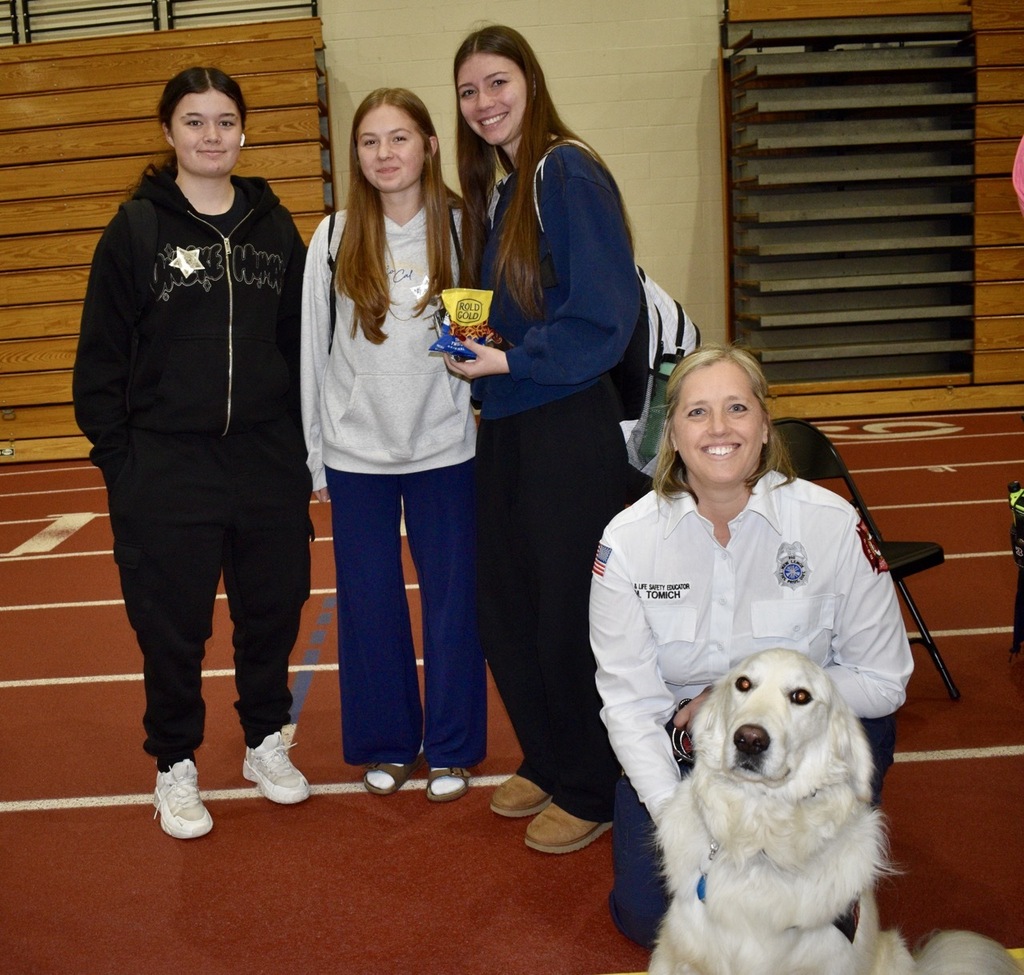 Four individuals stand on a red track. Two wear white jackets. Another wears a black hoodie. A dog stands nearby.