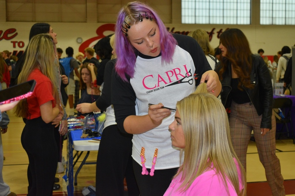 A girl with purple hair is doing another girl's hair at a school event. Other students are present.