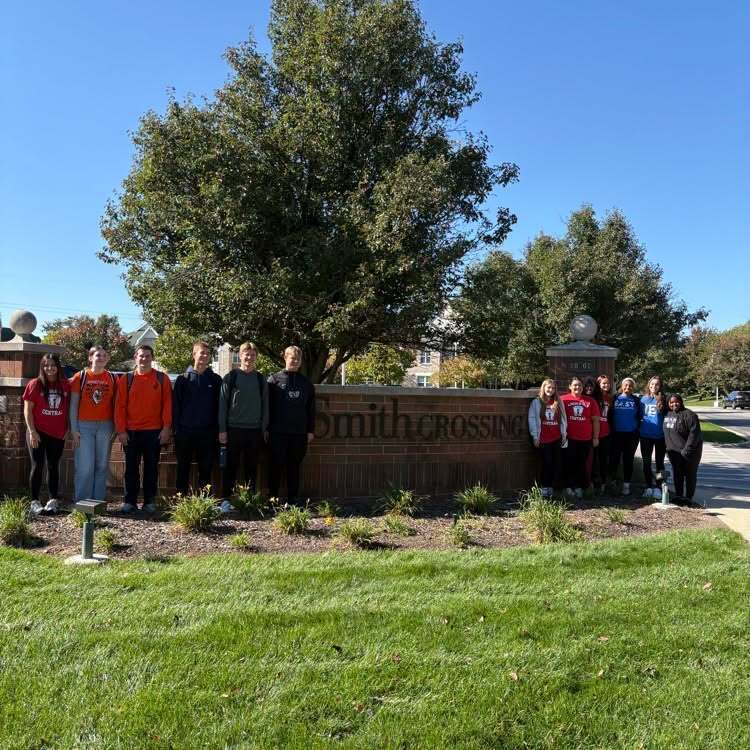 People stand in two rows in front of a school sign with a tree and fountain behind them.