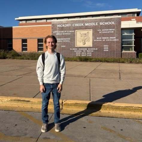 A student stands outside, wearing a white shirt, jeans, and sneakers. The building behind has a sign reading "Hickory Creek Middle School."