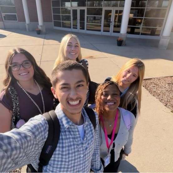 Five students stand in a row, smiling and posing for a photo on a school sidewalk.
