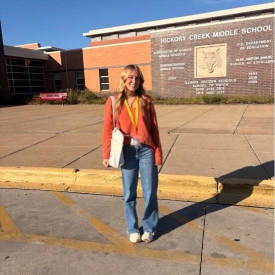 A person in front of a building with text. Person wears jeans and white shoes.