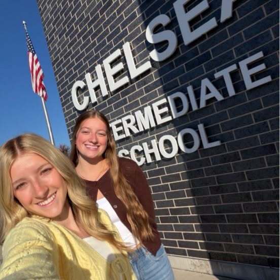 Two girls smiling at a building with brick walls and a sign that says "Chelsea Intermediate School".