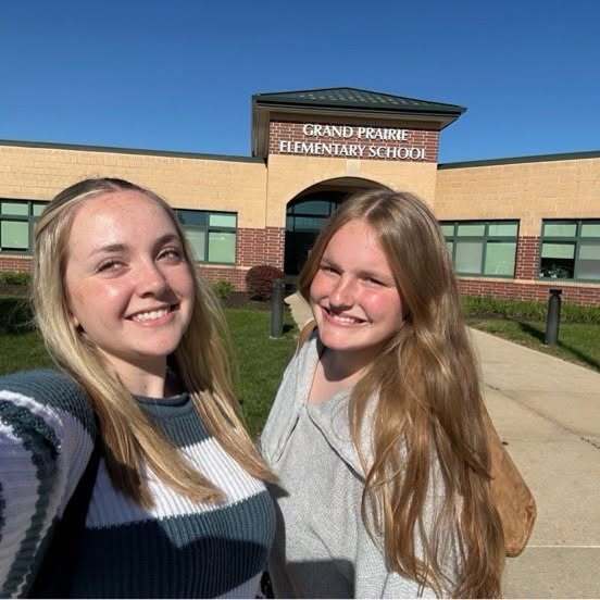 Two girls standing, smiling, outside a building with "Grand Prairie Elementary School" sign and green grass.