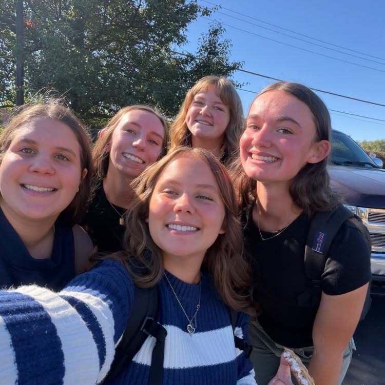Five girls smiling and posing for a selfie with a pickup truck in the background.
