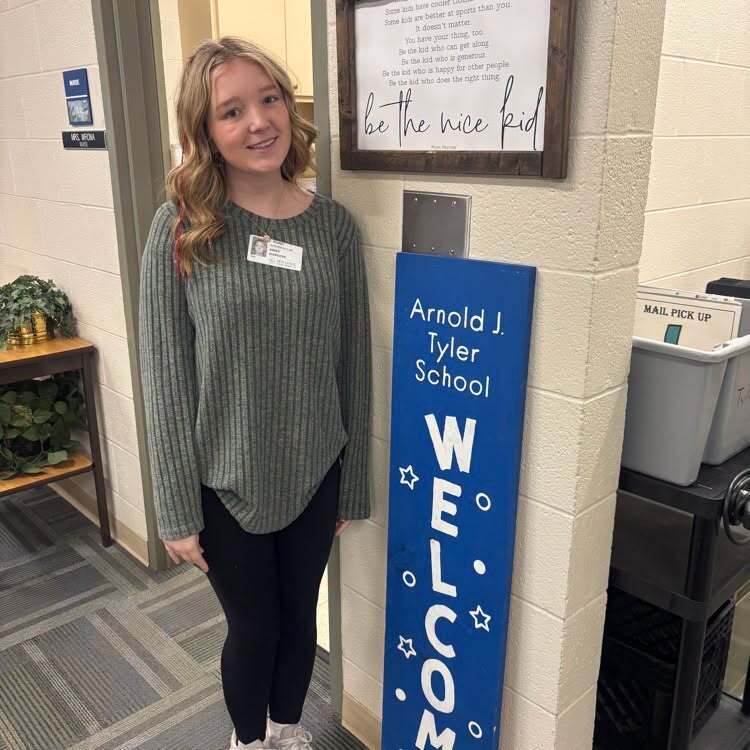 Teen girl in green top and black pants stands next to blue sign. Behind her is a sign on wall and potted plants.