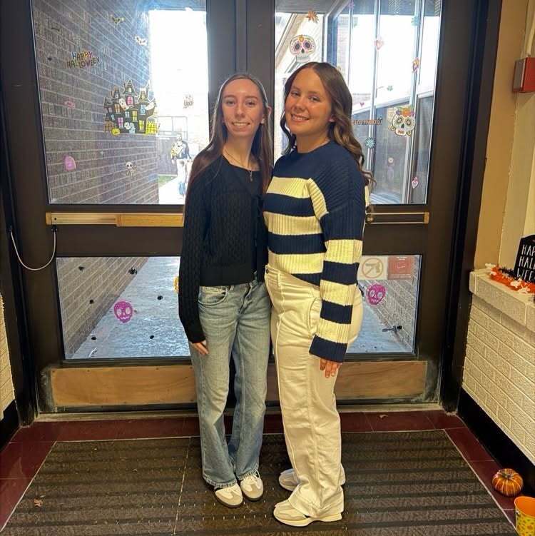 Two students stand by a door with pumpkins and decorations. Both wear white shoes. The one on the right has a sweater with horizontal stripes.