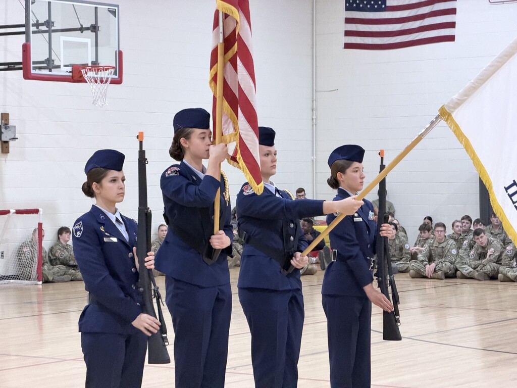 Four cadets hold flags and rifles, standing in a gym with a basketball hoop and seated cadets.