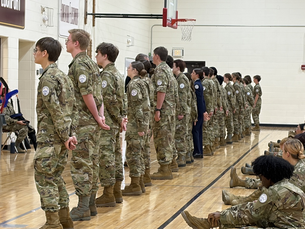 People in uniforms lined up on a gym floor with others sitting behind them.