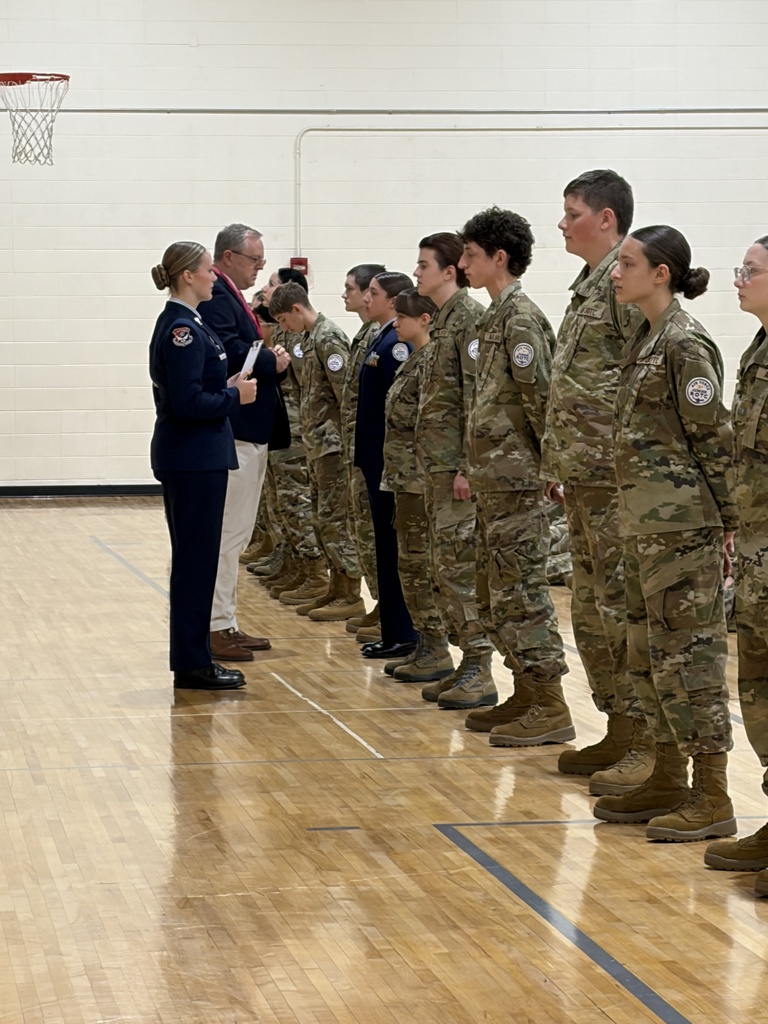 Military personnel stand in a line on a gym floor. A woman in uniform speaks to a man.
