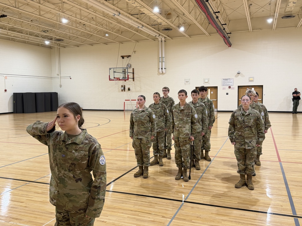 Group of students in military uniforms, saluting, stand on a basketball court. A person walks in the distance.