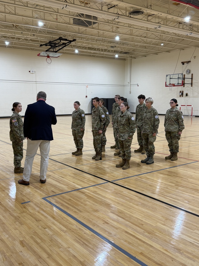 A man stands in front of a group of military-dressed students in a gymnasium.