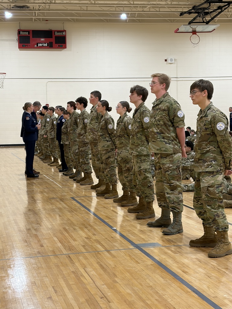 Group of students in uniforms standing in a line in a gym, a man speaks to them.
