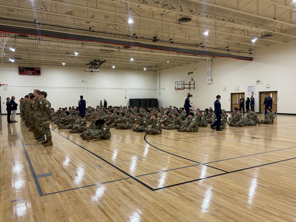 Several people in camouflage uniforms on a basketball court, some seated, others standing, with a basketball hoop in the background.