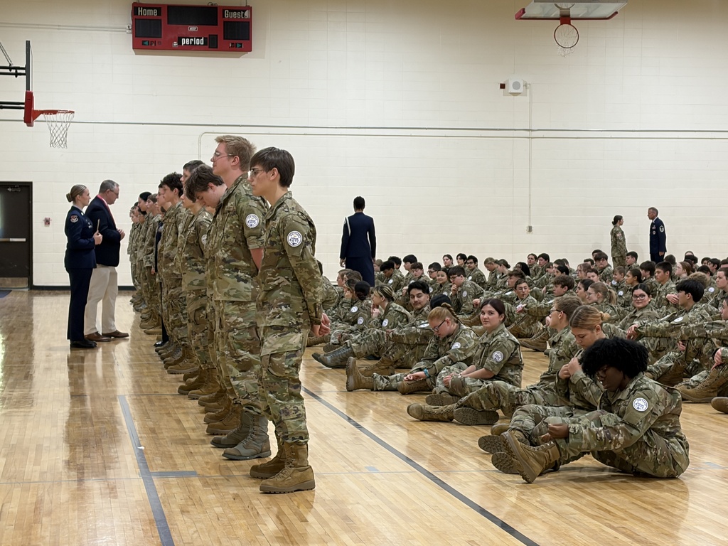 Group of uniformed people stand in lines and sit on floor in gym, a few people standing by wall