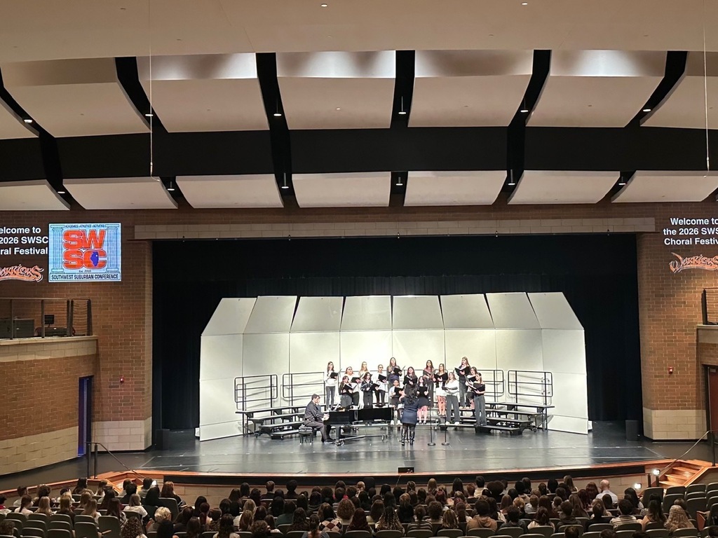 Choir performs on stage with audience seated in rows; stage backdrop is white, with brick walls on sides.